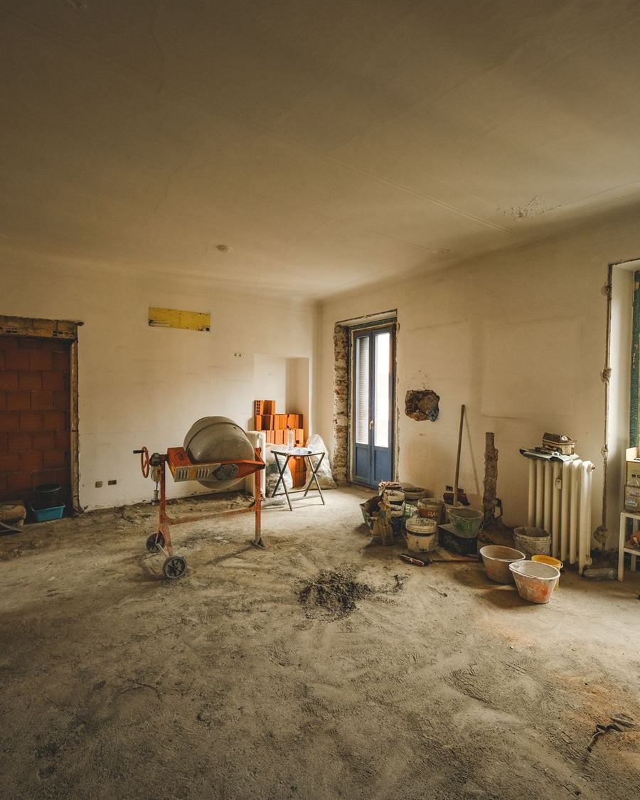 Divisão de casa antiga em remodelação, paredes picadas, escadote e baldes, luz a entrar pelas portas.