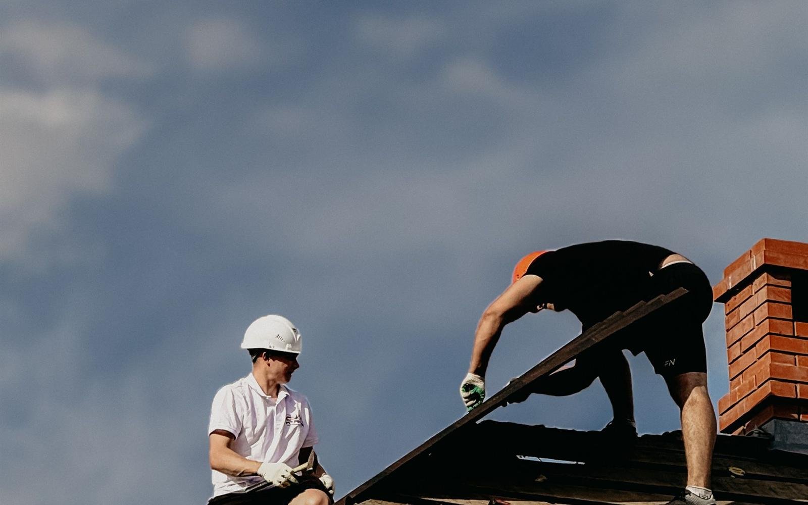 Dois trabalhadores a reparar o telhado de telha de uma moradia, sob céu azul